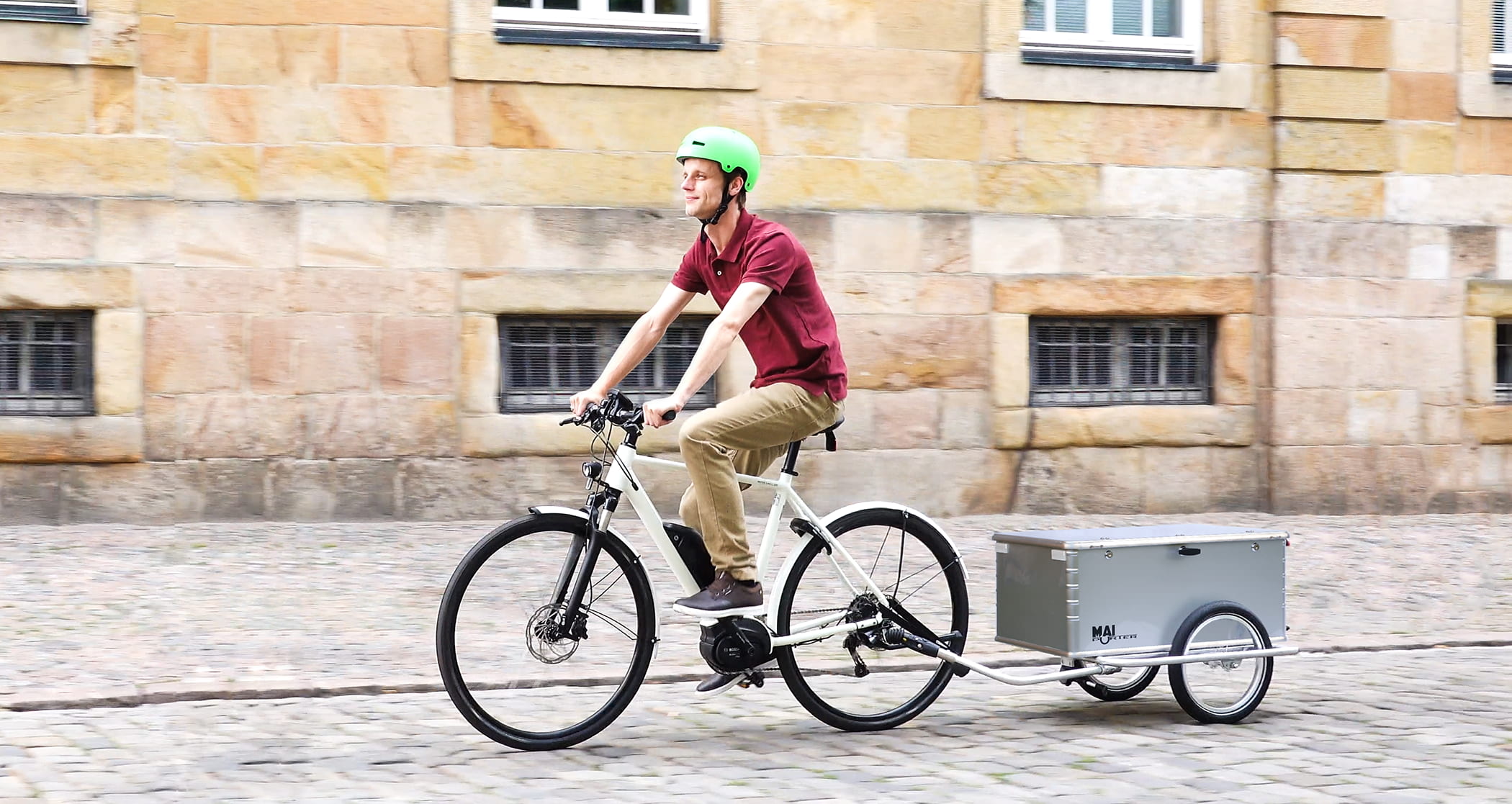Ein lächelnder Mann fährt mit einem E-Bike und zieht den MaiPorter Fahrradanhänger über unebenes Kopfsteinpflaster.
